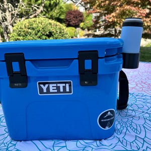 Op de afbeelding: Een blauwe YETI-koeler met zwarte grendels en een zwarte riem. Een wit YETI-logo staat op de voorkant. Een lichtblauwe en zwarte beker is aan de zijkant bevestigd. De koeler staat op een patroon deken.
