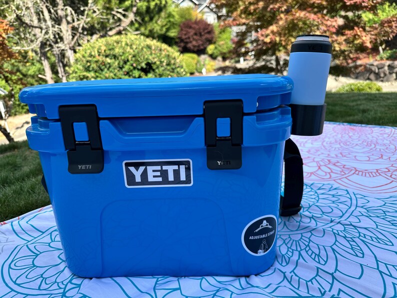 Op de afbeelding: Een blauwe YETI-koeler met zwarte grendels en een zwarte riem. Een wit YETI-logo staat op de voorkant. Een lichtblauwe en zwarte beker is aan de zijkant bevestigd. De koeler staat op een patroon deken.