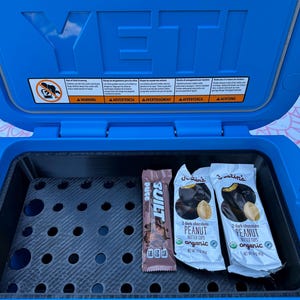 May include: A blue YETI cooler with the lid open, revealing a black interior with a grid. Inside are two Justin's peanut butter cup packages and a Built protein bar. The cooler has warning labels on the inside of the lid.