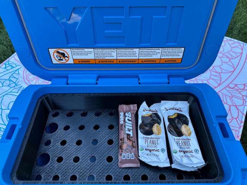 May include: A blue YETI cooler with the lid open, revealing a black interior with a grid. Inside are two Justin's peanut butter cup packages and a Built protein bar. The cooler has warning labels on the inside of the lid.