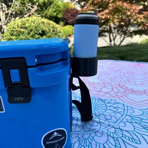 Op de afbeelding: Een blauwe koeler met een zwarte handgreep en een zwarte bekerhouder. Een lichtblauw blikje zit in de bekerhouder. Op de koeler staat het woord "YETI" en een sticker met de tekst "ADJUSTABLE STRAP". De koeler staat op een patroon deken.