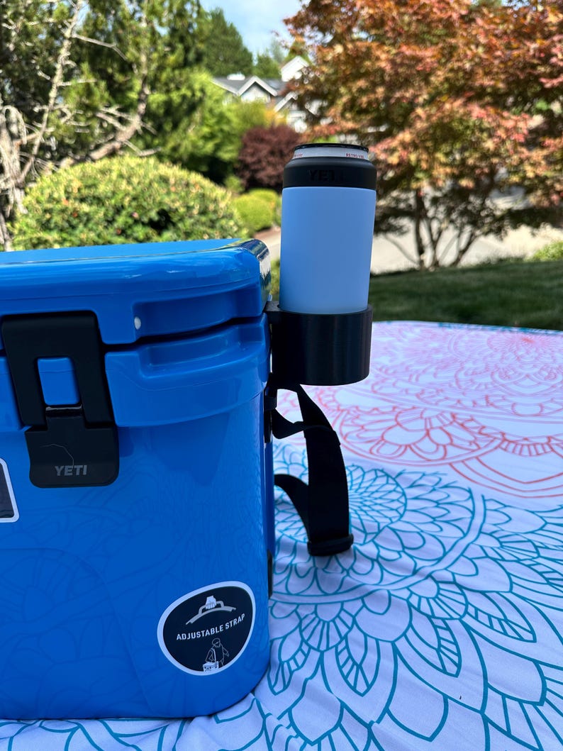 Op de afbeelding: Een blauwe koeler met een zwarte handgreep en een zwarte bekerhouder. Een lichtblauw blikje zit in de bekerhouder. Op de koeler staat het woord "YETI" en een sticker met de tekst "ADJUSTABLE STRAP". De koeler staat op een patroon deken.