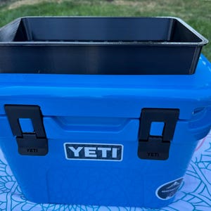 May include: A blue YETI cooler with black latches and a black tray on top. The cooler has the YETI logo on the front. The cooler is sitting on a patterned surface, with green grass in the background.