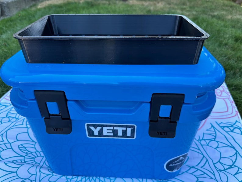 May include: A blue YETI cooler with black latches and a black tray on top. The cooler has the YETI logo on the front. The cooler is sitting on a patterned surface, with green grass in the background.