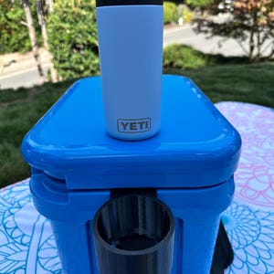 Op de afbeelding: Een lichtblauwe Yeti-blikisolator met een zwarte bovenkant, rustend op een blauwe koeler. Het Yeti-logo is zichtbaar op de voorkant. De koeler heeft een ingebouwde bekerhouder. De achtergrond toont groen gebladerte.