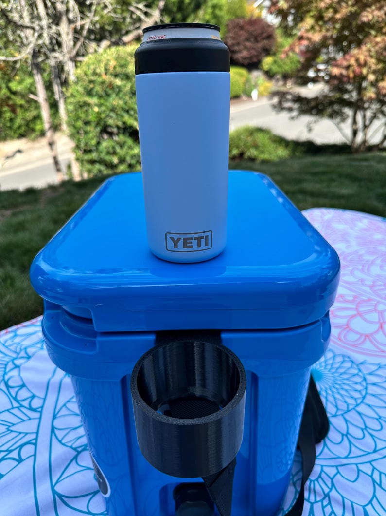 Op de afbeelding: Een lichtblauwe Yeti-blikisolator met een zwarte bovenkant, rustend op een blauwe koeler. Het Yeti-logo is zichtbaar op de voorkant. De koeler heeft een ingebouwde bekerhouder. De achtergrond toont groen gebladerte.