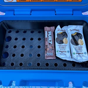 May include: A blue cooler with a black interior containing a Built protein bar and two Justin's dark chocolate peanut butter cups. The cooler has a perforated shelf and is open.