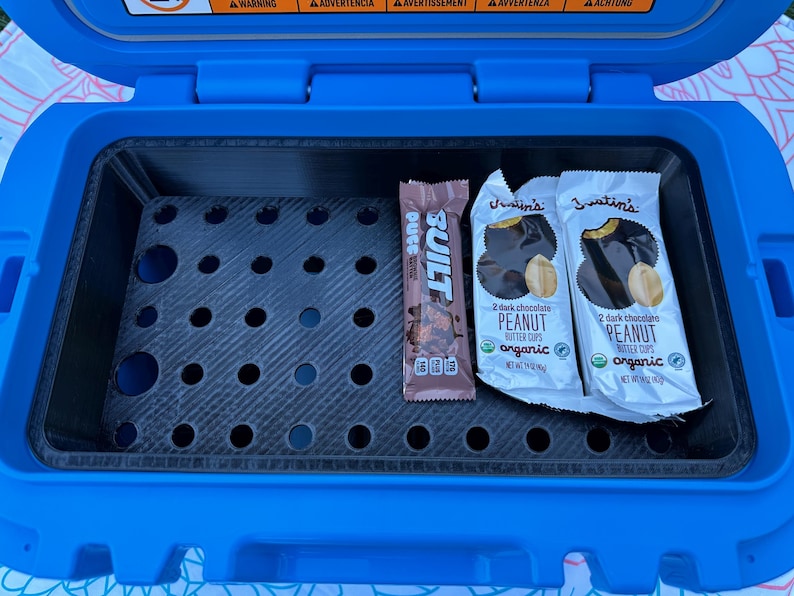 May include: A blue cooler with a black interior containing a Built protein bar and two Justin's dark chocolate peanut butter cups. The cooler has a perforated shelf and is open.