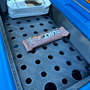 May include: A blue cooler with a black perforated tray holding two wrapped protein bars and a Built bar. The protein bars are white with the text "Peanut" and "Organic". The Built bar is pink and brown.