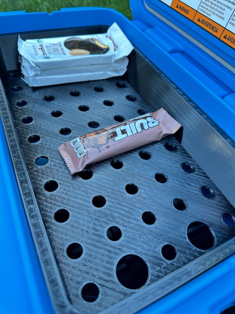 May include: A blue cooler with a black perforated tray holding two wrapped protein bars and a Built bar. The protein bars are white with the text "Peanut" and "Organic". The Built bar is pink and brown.