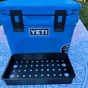 May include: A bright blue YETI cooler with black latches and the YETI logo. A black tray with multiple circular holes sits in front of the cooler. The cooler is on a patterned blanket, with green grass in the background.