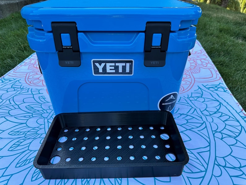 May include: A bright blue YETI cooler with black latches and the YETI logo. A black tray with multiple circular holes sits in front of the cooler. The cooler is on a patterned blanket, with green grass in the background.