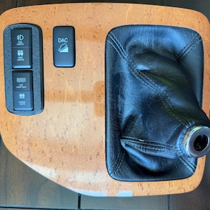 May include: A car interior panel with a wood grain effect. It features a black leather gear shift boot and several black buttons labelled "Fog Lights", "Spot Lights", "LED Light Bar", "Reverse Lights", and "DAC".