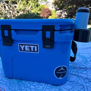 Op de afbeelding: Een blauwe YETI-koeler met zwarte grendels en een zwarte riem. Een blauwe beker en een zwarte bekerhouder zijn aan de zijkant bevestigd. De koeler staat buiten op een patroon deken.