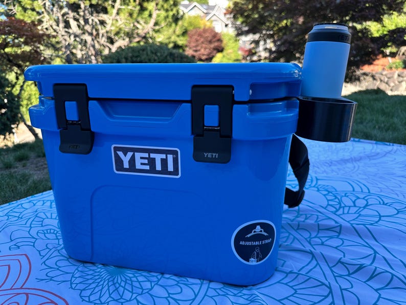 Op de afbeelding: Een blauwe YETI-koeler met zwarte grendels en een zwarte riem. Een blauwe beker en een zwarte bekerhouder zijn aan de zijkant bevestigd. De koeler staat buiten op een patroon deken.