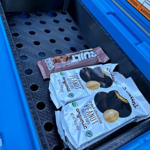 May include: A blue cooler interior with a black perforated tray holding two packages of Justin's organic dark chocolate peanut butter cups and a Built protein bar. The cooler has warning labels on the lid.