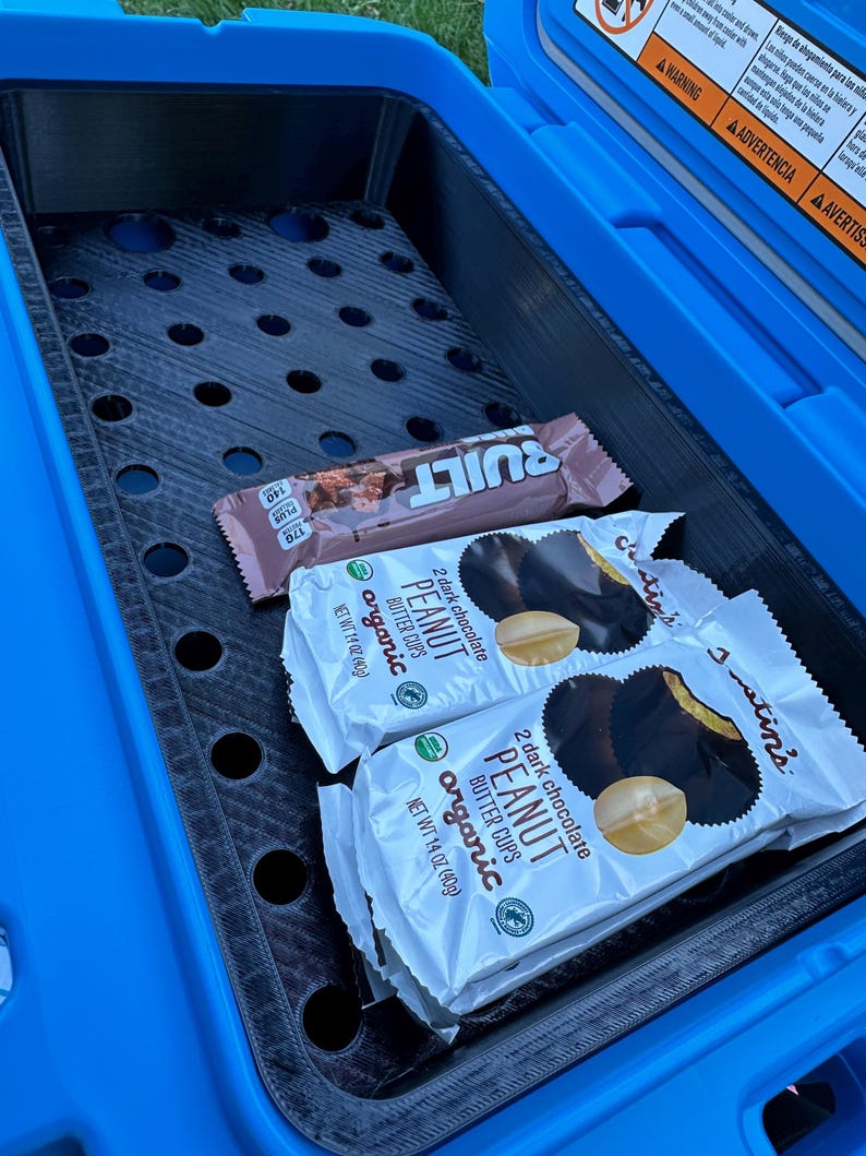 May include: A blue cooler interior with a black perforated tray holding two packages of Justin's organic dark chocolate peanut butter cups and a Built protein bar. The cooler has warning labels on the lid.