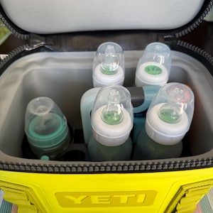 Puede incluir: Una nevera portátil YETI amarilla con cremallera superior, que contiene varios biberones con tetinas transparentes y tapas verdes y blancas. La nevera está abierta, revelando un interior gris. La nevera está sobre una superficie rayada.