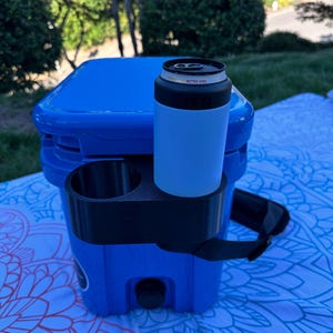 Op de afbeelding: Een blauwe koeler met een zwarte bekerhouder en een lichtblauwe blikjeskoeler. Op de blikjeskoeler staat "Retro Vibe". De koeler heeft een zwarte riem en staat op een patroon deken.