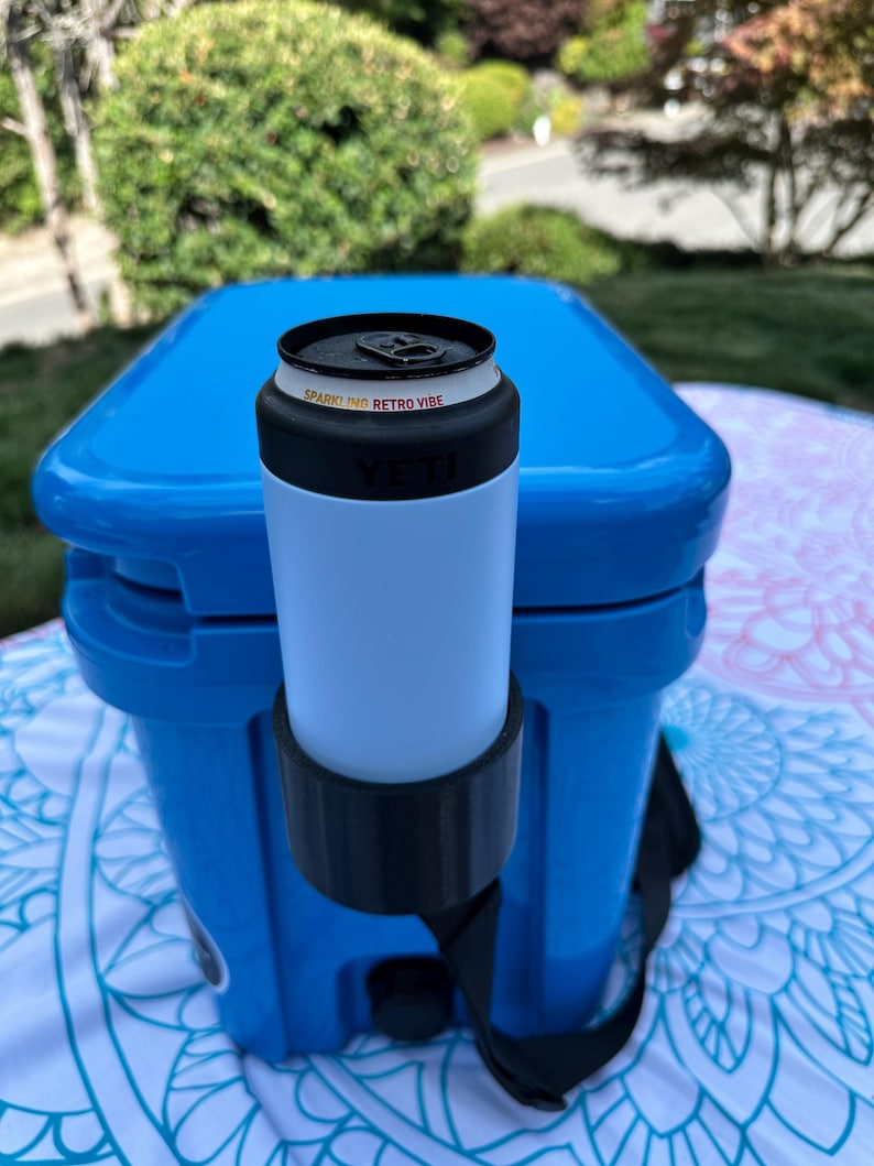 Op de afbeelding: Een blauwe koeler met een witte en zwarte drankhouder bevestigd. Een wit en zwart blikje met de tekst "SPARKLING RETRO VIBE" staat in de houder. De koeler staat op een patroon deken, met groen op de achtergrond.