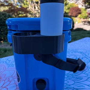 Op de afbeelding: Een blauwe koeler met een zwarte drankhouder bevestigd. Een lichtblauwe YETI-blikjeskoeler bevat een blikje met de tekst "RETRO VIBE". De koeler staat buiten op een patroonkleed.