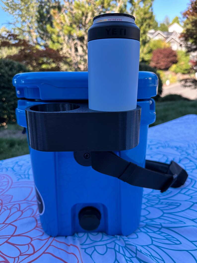 Op de afbeelding: Een blauwe koeler met een zwarte drankhouder bevestigd. Een lichtblauwe YETI-blikjeskoeler bevat een blikje met de tekst "RETRO VIBE". De koeler staat buiten op een patroonkleed.