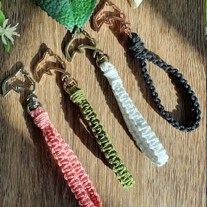 May include: Four macrame keychains in pink, green, white, and black. Each keychain features a woven cord loop and a metal clasp with a star-shaped design. The keychains are arranged on a wooden surface with floral accents.