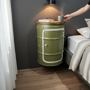 Peut inclure: Une table de chevet verte en forme de baril avec un dessus en bois et une porte bordée de blanc. La table est fixée à un mur gris foncé. Un livre et des lunettes sont posés dessus, et une personne tient une tasse.
