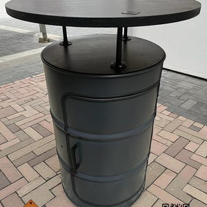 Könnte beinhalten: Ein schwarzer Tisch mit runder Tischplatte und einem fassförmigen Unterbau. Der Tisch hat eine dunkle Oberfläche und wird von Metallbeinen getragen. Der Unterbau hat eine Tür und einen Griff. Der Tisch ist im industriellen Stil gestaltet. Der Tisch hat einen Durchmesser von ca. 91 cm.