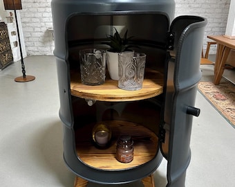 May include: A dark grey barrel-shaped cabinet with a wooden top and shelves. The cabinet door is open, revealing two shelves with decorative items. The cabinet is supported by wooden legs and has a handle. The cabinet has a "LOFT DESIGN" logo.