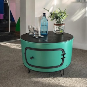 Puede incluir: Una mesa auxiliar de color verde azulado con una tapa negra y un solo cajón. La mesa tiene tres patas de horquilla y está sobre una alfombra gris claro. Hay una botella de ginebra, dos vasos y un jarrón con plantas verdes sobre la mesa.