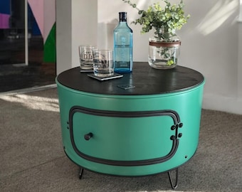 Puede incluir: Una mesa auxiliar de color verde azulado con una tapa negra y un solo cajón. La mesa tiene tres patas de horquilla y está sobre una alfombra gris claro. Hay una botella de ginebra, dos vasos y un jarrón con plantas verdes sobre la mesa.