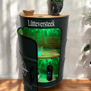 Peut inclure: Un meuble en forme de baril vert foncé avec un dessus et des étagères intérieures en bois. La porte de l'armoire est ouverte, révélant un intérieur éclairé en vert avec une bouteille de vin et un verre. Le texte "Lütteversteek" est affiché à l'avant.