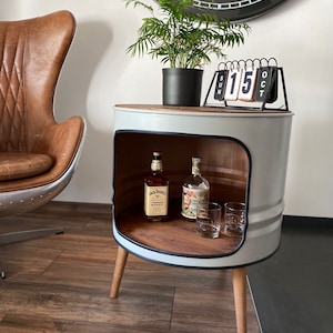 Op de afbeelding: Een grijze metalen vat-bijzettafel met een houten blad en zwarte leren afwerking. De tafel heeft een plank en staat op houten poten. Er staan twee flessen en twee glazen op de plank. Een potplant en een kalender staan op de tafel.