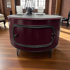 Può includere: Un tavolino da caffè a forma di tamburo, color bordeaux, con una porta a cerniera e finiture nere. Il tavolo ha un piano scuro e poggia su gambe a forcina nere. Due bicchieri con testo sono sul tavolo.