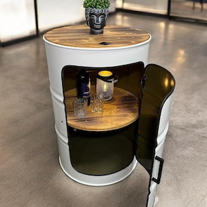 Puede incluir: Barril de metal blanco con tapa de madera y una puerta que se abre para revelar un estante. El barril está decorado con una pequeña estatua de Buda y tiene una pequeña vela en su interior.