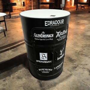 Può includere: Un mobiletto nero in metallo a forma di barile con un piano bianco. Il mobiletto è decorato con etichette di vari marchi di whisky scozzese, tra cui Edradour, The Glendronach, Ardbeg, Springbank, Glenfiddich, Bowmore e Laphroaig.