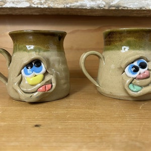 May include: Two ceramic mugs with brown and green glaze. Each mug has a face with blue eyes, a yellow nose, and a pink tongue. One mug has a green leaf on its cheek.