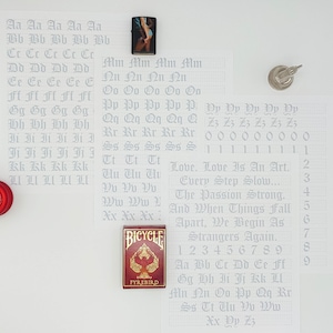 May include: A white sheet of paper with a grid pattern and handwritten letters of the alphabet in a gothic font. The letters are written in rows, with each row representing a different letter. The letters are written in a light gray color. The paper is surrounded by other objects, including a red yo-yo, a lighter, and a deck of playing cards.