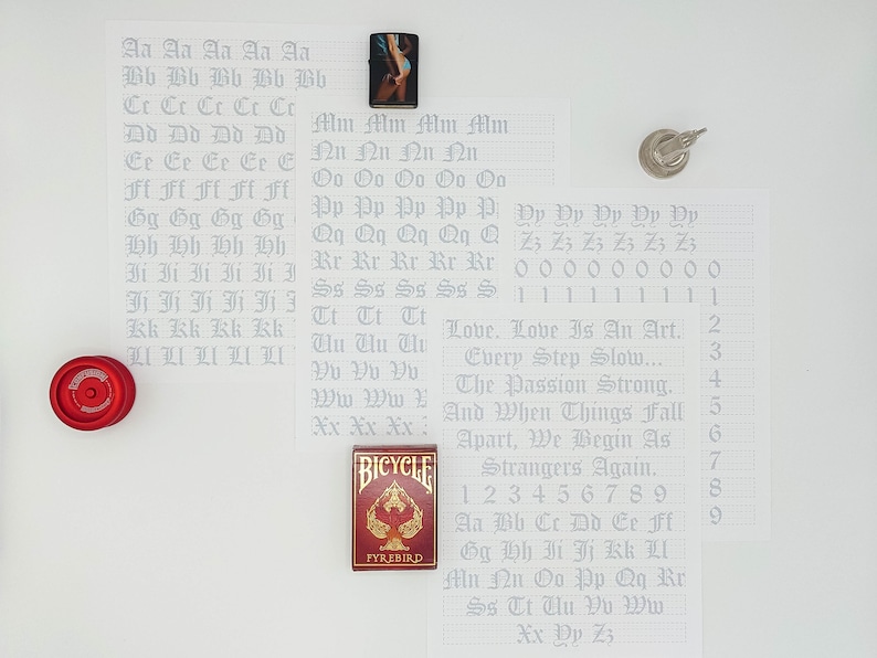 May include: A white sheet of paper with a grid pattern and handwritten letters of the alphabet in a gothic font. The letters are written in rows, with each row representing a different letter. The letters are written in a light gray color. The paper is surrounded by other objects, including a red yo-yo, a lighter, and a deck of playing cards.
