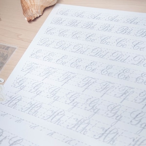 May include: A white sheet of paper with cursive letters of the alphabet written in grey ink. The letters are written in rows, with each row containing a different letter. The letters are spaced evenly apart and are written in a neat and legible style.