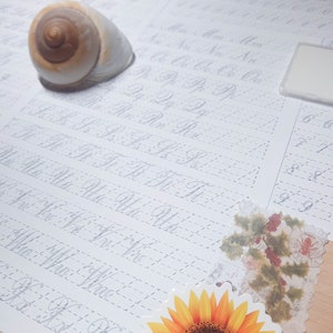 May include: A white sheet of paper with grey dotted lines and cursive handwriting practice. The paper is on a wooden surface with a seashell, a sunflower sticker, a floral sticker, and a black pen.