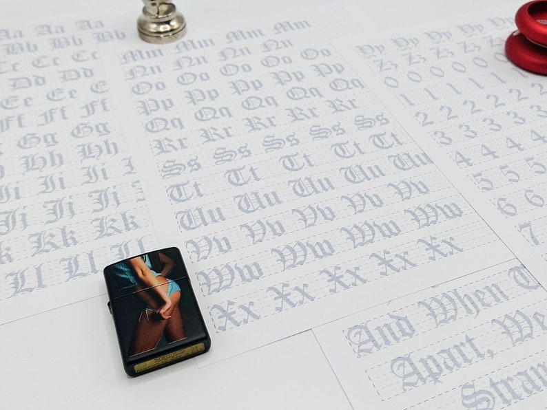 May include: A black lighter with a colorful image of a woman in a blue bikini. The lighter is on a white surface with a sheet of paper with the alphabet written in cursive.