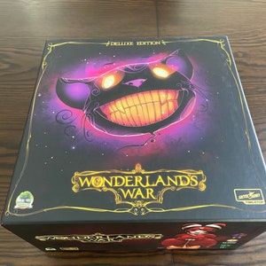 Puede incluir: Una caja negra con un borde dorado y el texto "Wonderlands War Deluxe Edition" en la parte delantera. La caja presenta una ilustración morada y negra de un gato con ojos y dientes amarillos brillantes. El texto "Wonderlands War" está en letras doradas en la parte delantera de la caja.