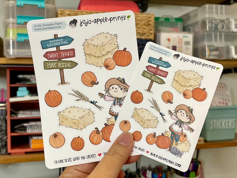 Op de afbeelding: Twee witte stickervellen met jojo-apple-prints branding, een vogelverschrikker, hooibalen, pompoenen, korenaren en wegwijzers met de tekst Pumpkin Decorating, Sweet Treats en Hay Ride. De tekst luidt I'd love to see what you create!