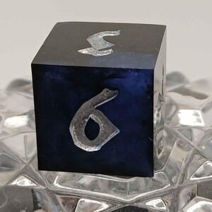 Pode incluir: Um cubo preto com numerais prateados. O número seis é visível em um lado. O cubo tem um interior azul escuro. O cubo está sobre uma superfície clara e texturizada.