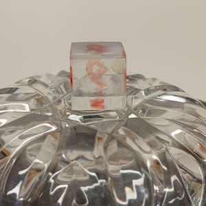 Op de afbeelding: Een helder, vierkant hars object met een rood, abstract ontwerp aan de binnenkant, staat op een heldere, facetgeslepen glazen kom. De kom heeft een herhalend geometrisch patroon. Het hars object lijkt een deksel of stopper te zijn.