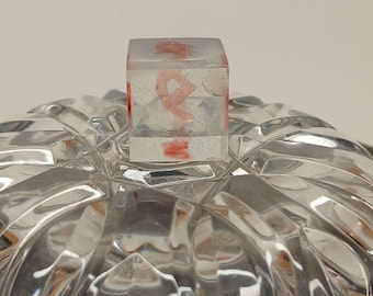 Könnte beinhalten: Ein klarer, quadratischer Gegenstand aus Harz mit einem roten, abstrakten Design im Inneren sitzt auf einer klaren, facettierten Glasschale. Die Schale hat ein sich wiederholendes geometrisches Muster. Der Harzgegenstand scheint ein Deckel oder ein Verschluss zu sein.