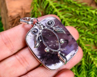 Puede incluir: Un colgante rectangular con una piedra pulida de color morado y blanco con inclusiones dendríticas oscuras. La piedra está engastada en un marco decorativo de plata de ley con detalles en forma de hoja y una anilla marcada con 925.