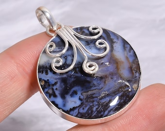 Puede incluir: Un colgante redondo con una piedra pulida de varios colores con patrones en espiral de azul oscuro, negro y blanco. La piedra está engastada en un marco de plata con un adorno de alambre de plata en espiral en la parte superior y una anilla.
